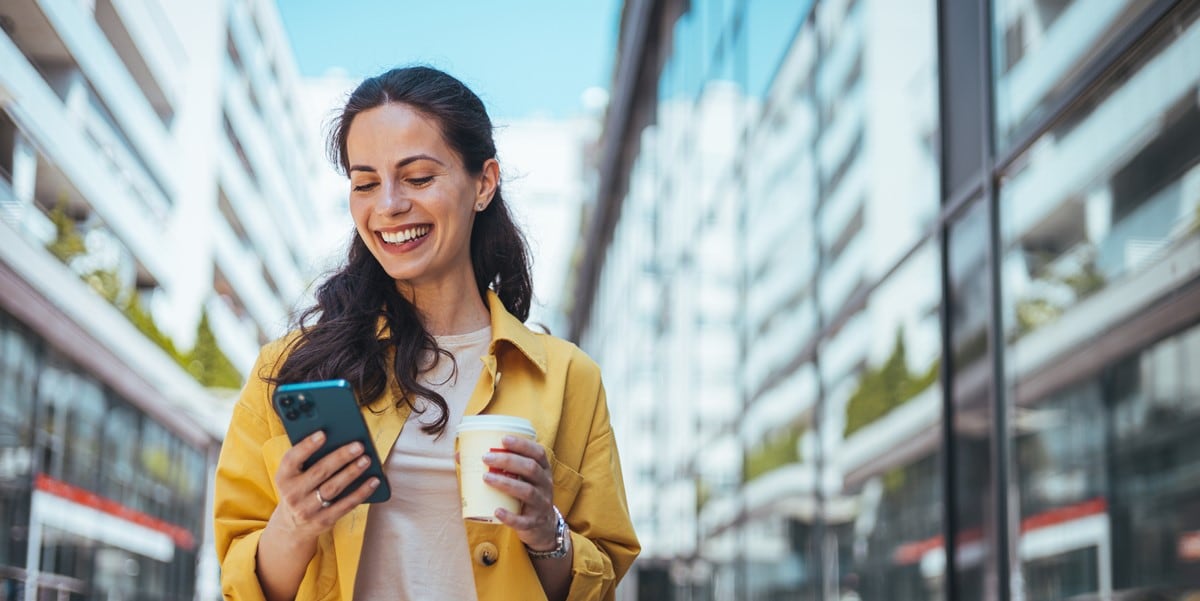 woman outdoors in city looking at phone with coffee cup in hand